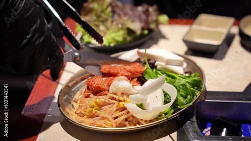 Korean BBQ Side Dishes (Banchan) Preparation,  Kimchi, Bean Sprouts, and Onion on Plate, Hand Using Tongs to Arrange Fresh Vegetables and Kimchi for Grilling on K-BBQ Table