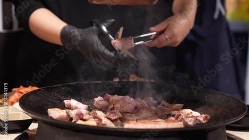 Authentic Korean Food Experience, Close-up of Hand Wearing Black Glove Grilling Samgyeopsal (Pork Belly) with Steam, Chef Using Tongs to Flip Pork Cut on Flat Top Grill