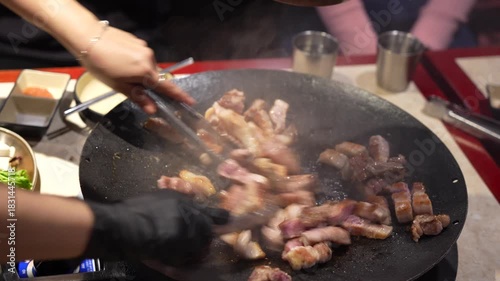 Authentic Korean Food Experience, Korean Samgyeopsal (Pork Belly) Sizzling on a Hot Cast Iron Griddle in Restaurant, Close-up of Hand Using Tongs to Cook Sliced Pork on Flat Top Korean BBQ