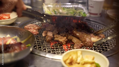 Close-up of Short Ribs (Galbi) Grilling on Charcoal Fire, Hand Using Tongs to Flip Marinated Meat on a Round Grill, Sizzling Meat Served with Side Dishes (Banchan) at a Restaurant