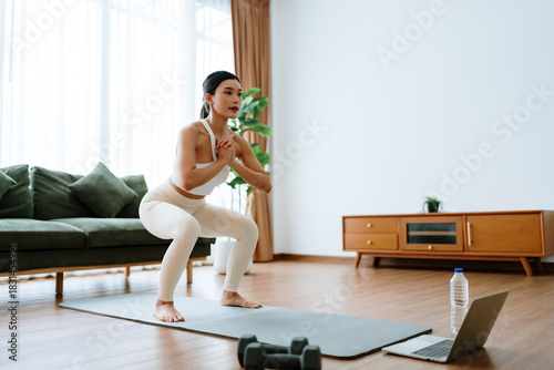 Young Asian beautiful woman doing aerobic exercise at home. Attractive girl doing  workout by squatting, follow instructions video from online trainer for health in house