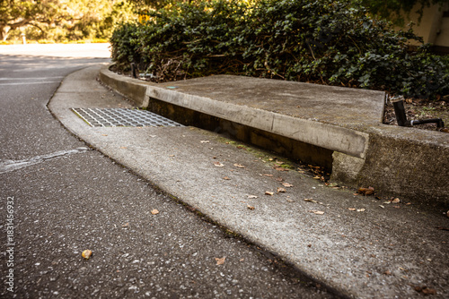 Fotografía Close-up view of a roadside storm drain and concrete curb on an asphalt street, with scattered autumn leaves and soft natural light