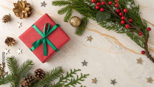 A red gift box with a green ribbon sits on a marble surface. Surrounding it are pine cones, evergreen branches, and festive decorations, creating a joyful holiday atmosphere.