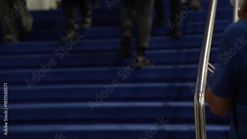 People walking up blue carpeted event stairs with stainless steel railings inside indoor venue, close-up view of legs, shoes and movement during business conference arrival