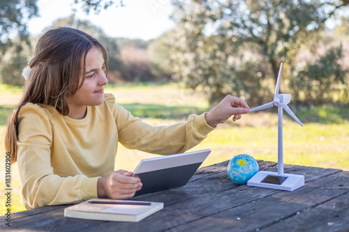 Young girl engaging with a miniature wind turbine model and a tablet, exploring concepts of renewable energy and environmental education in a natural outdoor setting on a wooden table
