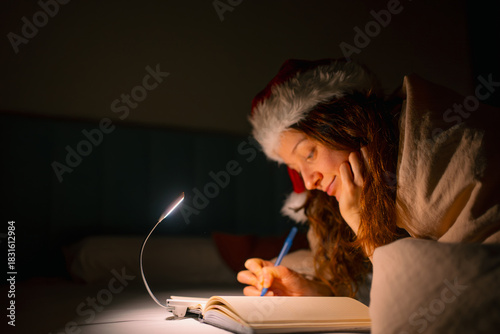 Woman wearing a warm santa hat and blanket writing in a notebook by the light of a book lamp, expressing holiday wishes during a cozy christmas season evening