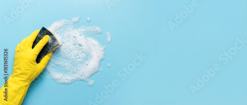 Hand in yellow rubber glove scrubbing surface with sponge, creating foam on blue background, showcasing cleaning process and hygiene practices