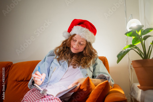 Young woman wearing a santa hat, sitting comfortably on a vibrant orange sofa, writing in a notebook, creating a christmas list or organizing holiday plans in a cozy home setting