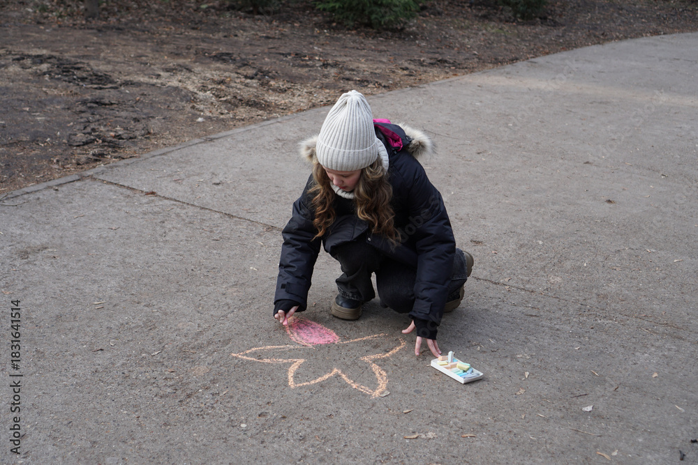 Fototapeta premium Child in a warm hat and jacket drawing a chalk flower on an asphalt pavement outdoors.