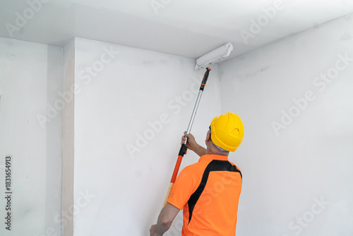 A construction worker paints a ceiling with a wide, specialized paint roller. Apartment renovation.