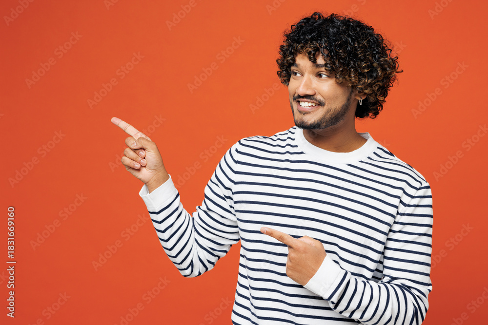 © ViDi Studio - Young smiling happy fun Indian man he wears striped sweatshirt casual clothes point index finger aside on empty blank area isolated on plain red orange background studio portrait. Lifestyle concept.