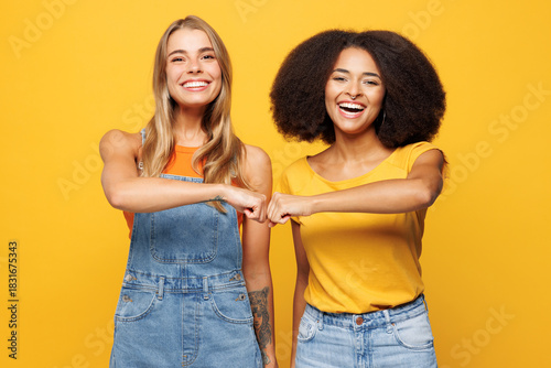 Two young smiling happy cheerful friends buddies women wear denim casual clothes together do fist bump gesture look camera isolated on plain yellow orange background studio portrait Lifestyle concept