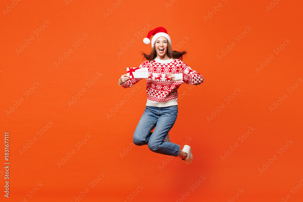 Obraz premium Full body merry young woman in red Christmas sweater Santa hat posing jump high hold gift coupon voucher card for store isolated on plain orange background Happy New Year celebration holiday concept