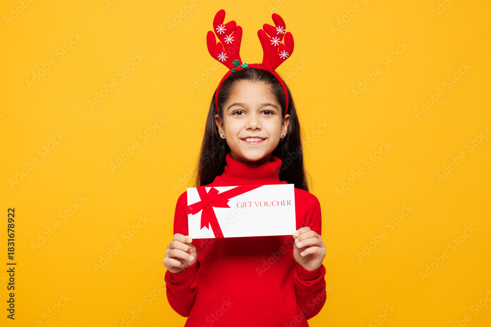 Fototapeta premium Merry little girl wear red clothes decorative deer horns on head posing hold store gift coupon voucher card isolated on plain yellow background. Happy New Year Christmas celebration holiday concept.