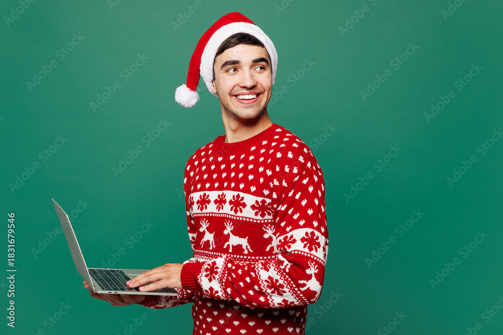 Naklejka premium Merry young IT man wear red warm Christmas sweater Santa Claus hat posing hold use work on laptop pc computer look aside isolated on plain green background. Happy New Year celebration holiday concept.