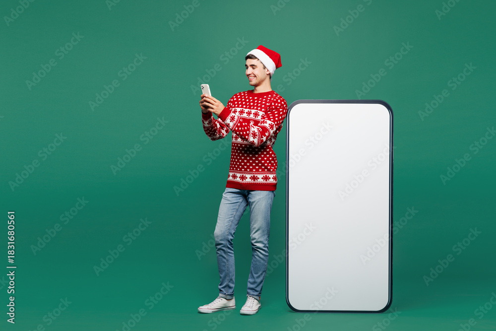 Fototapeta premium Full body merry young man in red Christmas sweater Santa Claus hat posing big huge blank screen area use mobile cell phone smartphone device isolated on plain green background Happy New Year concept