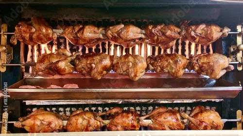 Rows of whole chickens roasting on a traditional rotisserie spit. Golden brown skin crisping over open flames at a market or butcher shop. Delicious street food cooking
