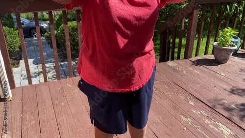 Video of an elderly woman standing outdoors on a brown painted deck, shown from torso to hips, wearing black pants and a red short-sleeve shirt while performing arm raise exercises with a cane on a su