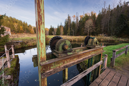 Принти на полотні Rusted metal gears and old sluice gate mechanism on a tranquil forest river in the countryside during autumn