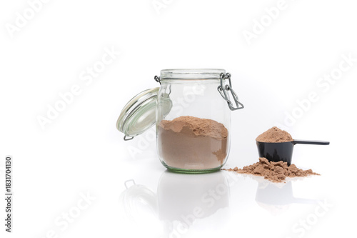 Horizontal image of a transparent jar of chocolate whey protein next to a scoop. White background with a reflective surface.