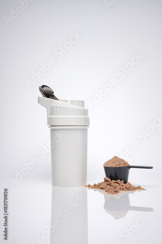 Vertical image of a shaker cup with whey protein in a scoop next to it. Light gray background with a reflective surface.