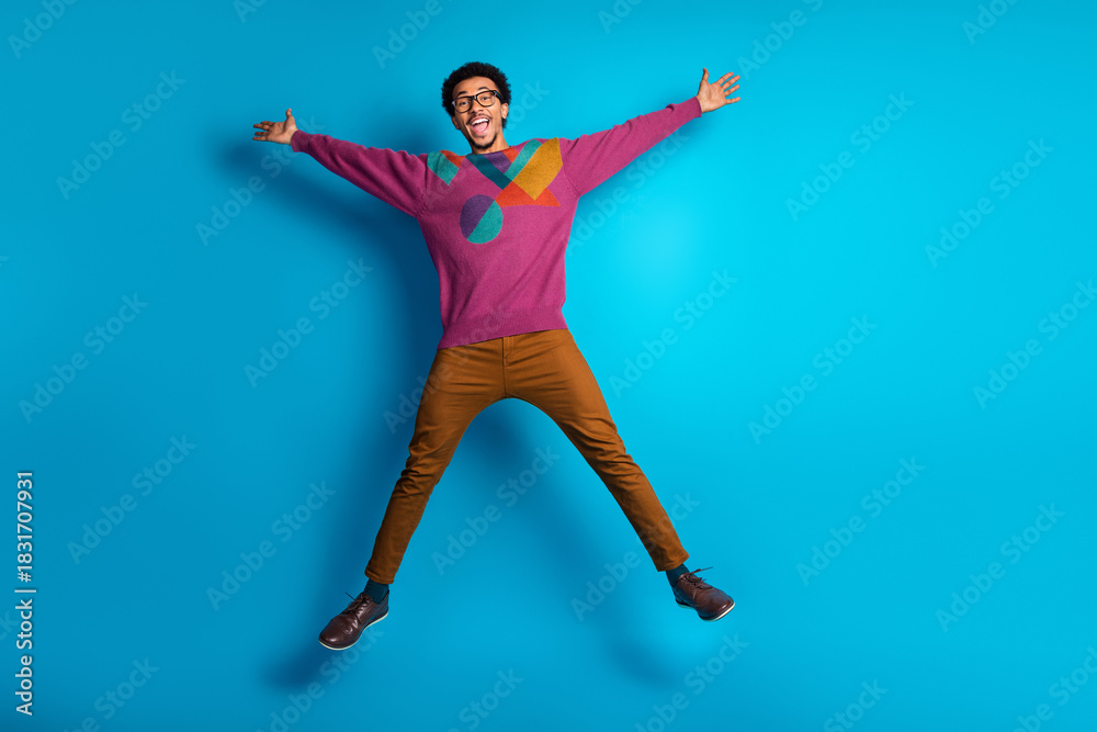 Obraz premium Excited young man jumping with joy in colorful casual outfit against a bright blue background promoting fun and lifestyle.
