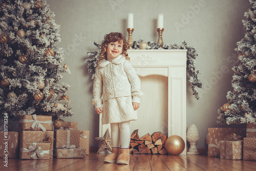 Hübsches kleines Mädchen mit Locken steht vor einem Kamin neben einem Weihnachtsbaum zu Weihnachten bei Abendstimmung mit Lichtern, Var. 1