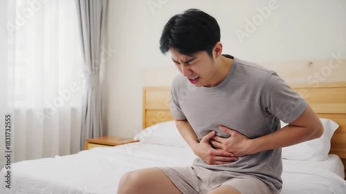 Young man in gray shirt sits on bed, visibly distressed from abdominal pain, with gradual camera zoom in, highlighting emotional struggle in a calm bedroom atmosphere