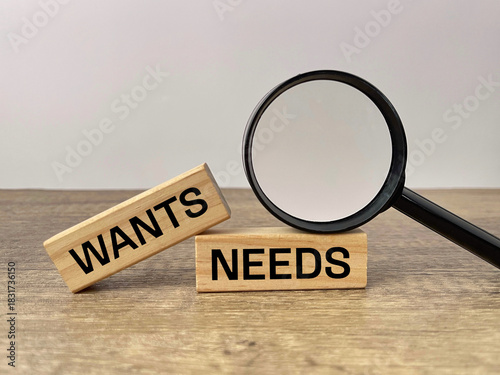 Wants vs Needs concept. WANTS and NEEDS written on wooden blocks with magnifying glass illustrating financial priorities and decision making.