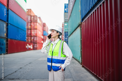 A female staff member reviews shipping details on the phone, standing between stacked cargo containers with a focused expression.