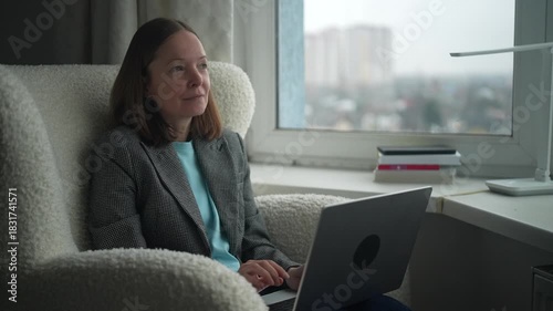 A woman sits comfortably in a soft chair, engaged with her laptop while a city skyline fills the background. Natural light creates a warm atmosphere as she works.