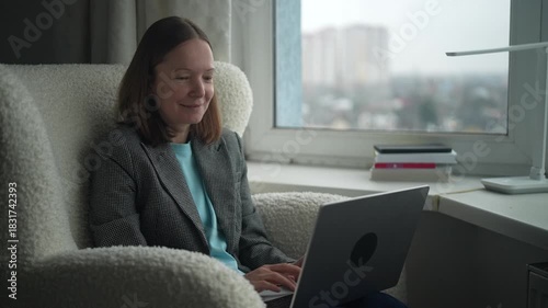 A woman sits comfortably in a chair, happily working on her laptop in a bright apartment. The city skyline can be seen through the window as she smiles.