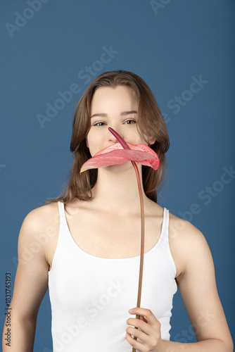 Beauty, make-up, style concept. Beautiful woman covering her mouth with pink Anthurium flower blossom. Model wearing white shirt and standing against blue background