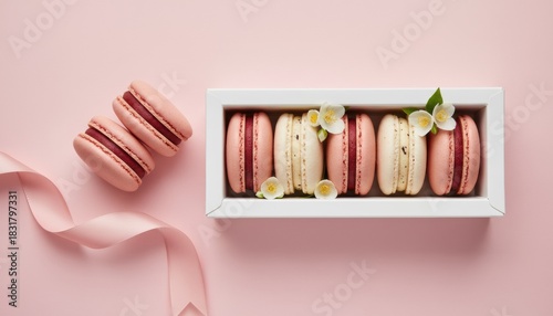 Pink and cream almond meringue macaron cookies in a white gift box with fresh jasmine flowers and a satin ribbon on a pastel pink background, top view flat lay composition