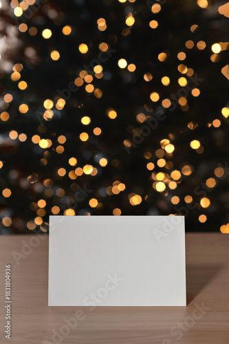 Festive greeting postcard mockup, blank paper card on table, blurred Christmas golden shining lights bokeh on background