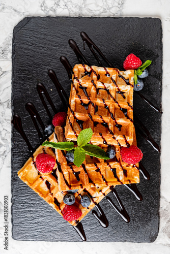 Two golden waffles are beautifully decorated with berries and chocolate on a sleek black slate