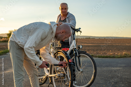 Two men with bicycles on rural road sunset, one fixing tire while other watches, teamwork and patience friends help, capturing companionship, active lifestyle, golden hour outdoor adventure