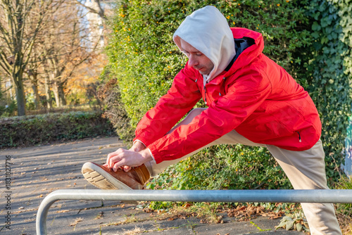 young man in red jacket stretching leg on park railing, warmup before run, solo training in cold air, discipline and small daily victories, public equipment for fitness routine, flexibility outdoors