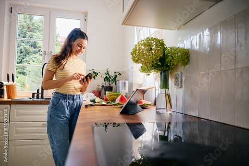 Young woman in kitchen checking recipe on her phone while preparing meal