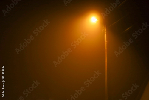 A bright orange streetlight on a tall pole pierces through thick fog or haze on a dark night. The light creates a halo, giving the scene a mystical and enigmatic appearance.