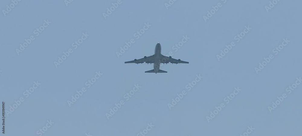 Fototapeta premium Wide-body Boeing 747 aircraft flying against a clear blue sky. Viewed from below, the image highlights the aircraft’s undercarriage, wings, and engines,ideal for illustrating aviation, aerospace.