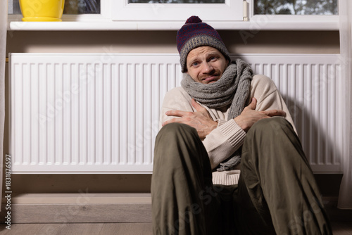 freezing man with hat and scarf in front of radiator in cold room at home. heating problems