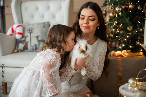 Special moments shared between a girl and her mother while holding a small rabbit near a beautifully decorated Christmas tree