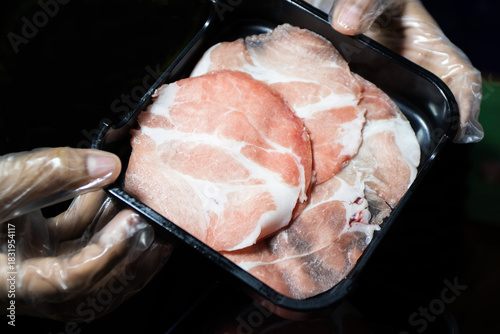 Close-up of raw, thinly sliced marbled meat (pork or beef neck) on a black tray, held by gloved hands, ready for cooking hot pot or yakiniku.