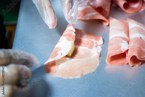 Gloved hand holding a piece of thinly sliced raw meat (pork or beef) with a small, yellow cube of cheese placed in the center, ready for preparation.