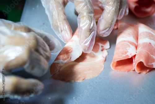 Gloved hand holding a piece of thinly sliced raw meat (pork or beef) with a small, yellow cube of cheese placed in the center, ready for preparation.