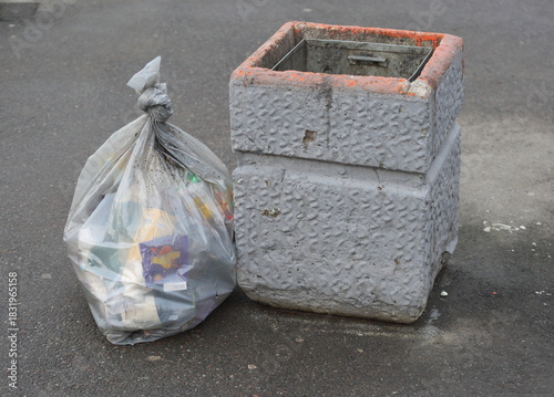 An empty concrete trash can and a knotted plastic bag of trash are sitting on the pavement