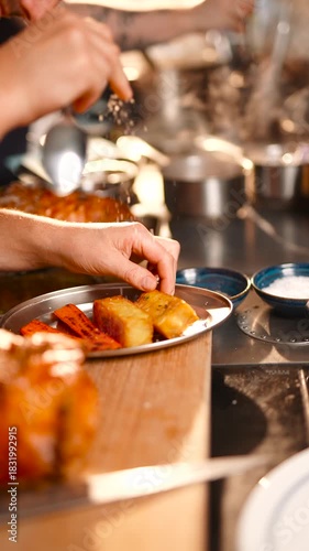 A person is cutting a piece of meat on a wooden cutting board. The meat is brown and has a crispy texture. A spoon is nearby, and there are potatoes on a plate. Concept of warmth and comfort