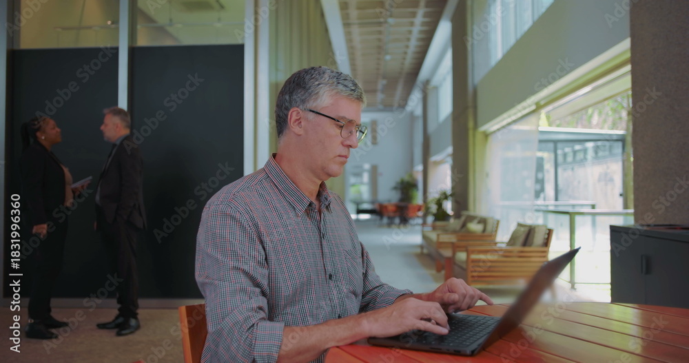 Fototapeta premium Middle aged man resuming work on laptop at shared office space, focused and typing with coworkers talking in background, overcoming concerns and staying productive