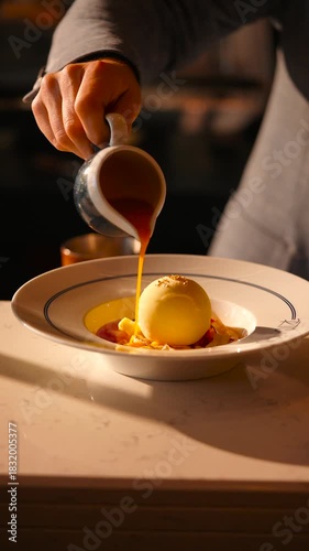 A person is pouring a liquid into a bowl of ice cream. The bowl is on a table and the person is wearing a blue shirt